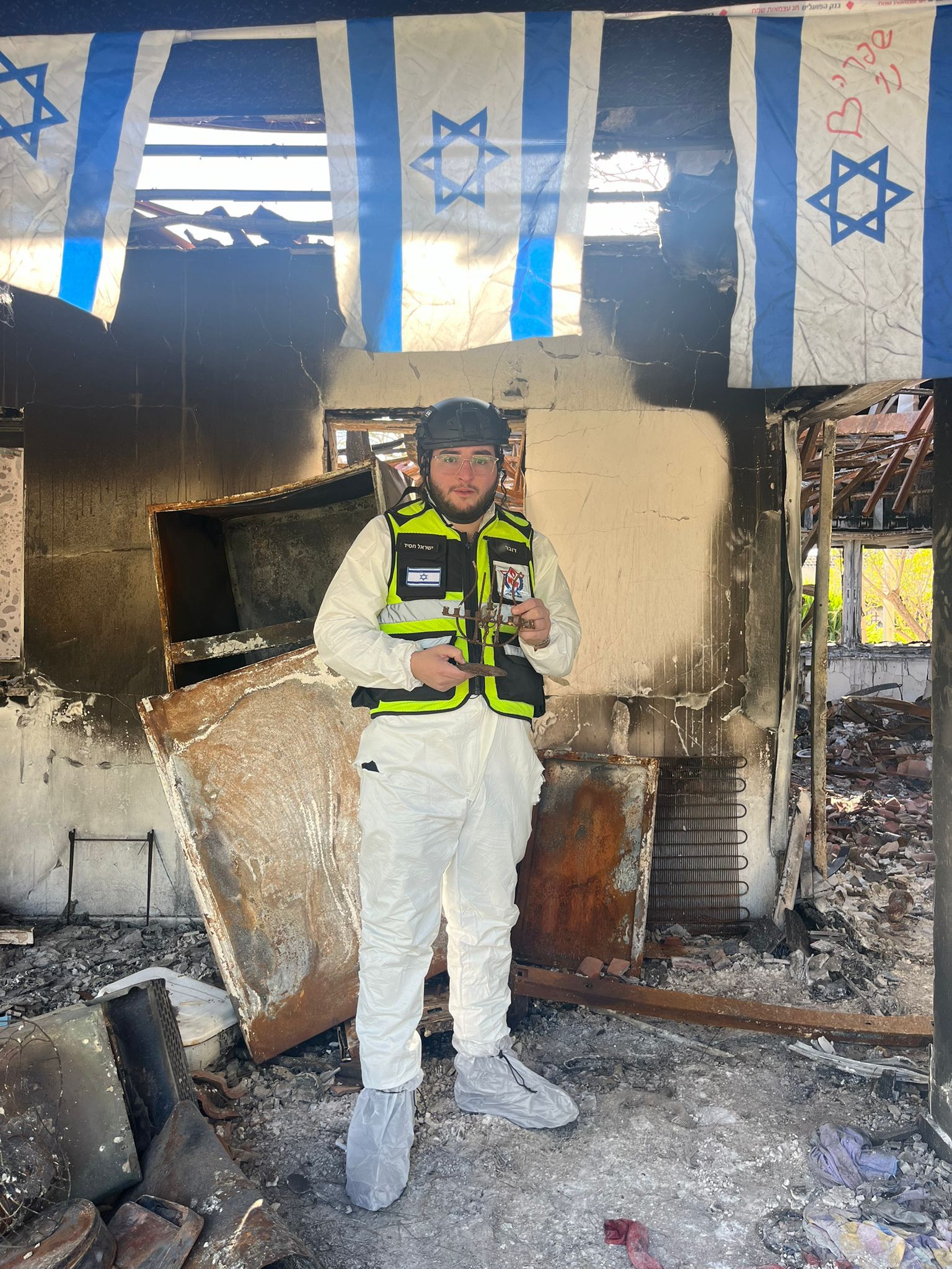 A lone ZAKA volunteer amid the ruins of a burned home, Israeli flags strung overhead.